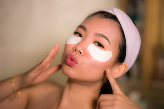 portrait of young happy and beautiful Asian Korean woman applying facial anti panda eyes patch at bathroom mirror in skin care and beauty treatment concept