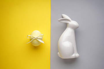 Easter bunny ceramic with yellow easter egg with ribbon on a yellow-grey background, top view.