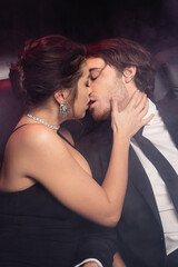 sensual woman in slip dress kissing man in suit on black with smoke.