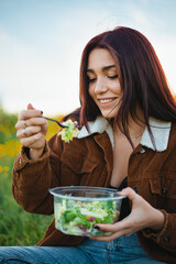 Happy teenager girl eating a salad while sitting on grass at a hill. She is enjoying the calm.