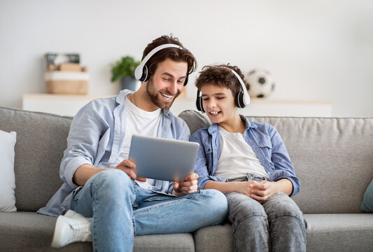 Modern father and son watching videos on digital tablet, wearing wireless headphones and smiling, surfing internet