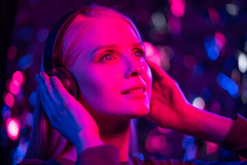 Happy young blonde woman listens to music with headphones in a nightclub. The girl smiles, dances and holds headphones with her hands. Blurred background, close-up, blue and red lights. Nightlife, fun