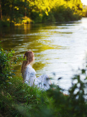 Cute beautiful young woman in white dress sitting on the river bank, sunny summer evening