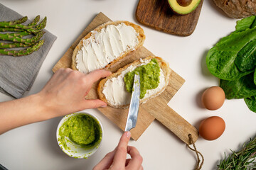 Female hand spreading mashed avocado on toasts with cream cheese on wooden cutting board. Green asparagus, egg, fresh salad, rosemary on white background. Top view, flat lay. Instruction, step 4