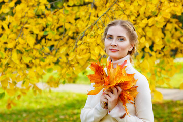 A beautiful young woman in an autumn park with a bouquet of maple leaves on a yellow tree background. CopySpace