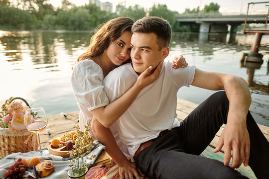 A young couple in love had a romantic picnic by the river at sunset. The girl hugs the young man and looks into the frame.