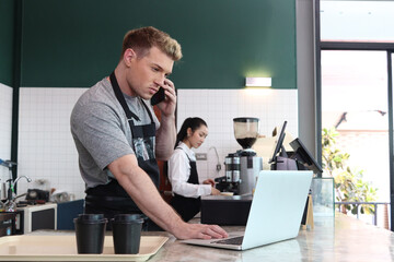 Young handsom male barista wearing apron, talking order on mobile phone and laptop computer while standing behind coffee shop counter bar, waiter staff working at cafe.