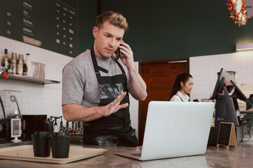 Young handsom male barista wearing apron, talking order on mobile phone and laptop computer while standing behind coffee shop counter bar, waiter staff working at cafe.