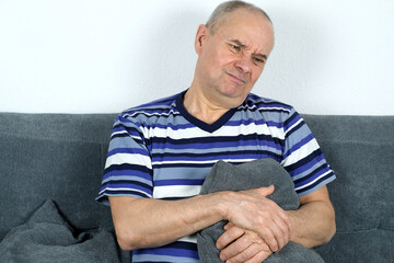 old bald man sitting on the sofa, pressing a pillow to his stomach, face grimace of suffering, stomach pain concept, gastritis, nausea