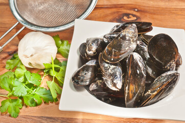 top view, close distance of a square, white bowl, filled with steamed mussels, a metal sieve, parsley and garlic head, on a wood cutting board
