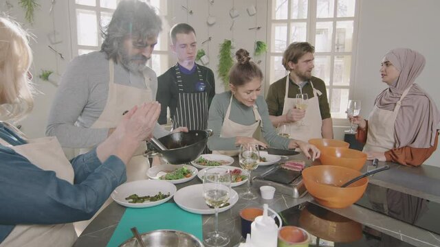 Slow-motion medium shot of small diverse group of people spending good time during cooking class with young successful male chef, talking and drinking wine while cooking