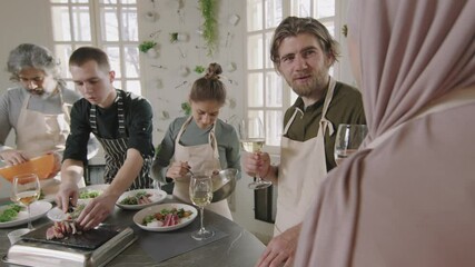 Slow-motion medium shot of diverse group of people talking and sipping wine while having cooking master class with chef in modern culinary school - Powered by Adobe