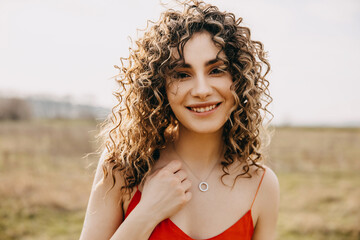 Young woman with curly hair smiling, outdoors, in a field at sunset, looking at camera.