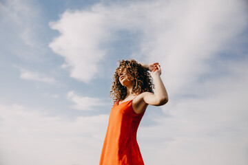 Young woman with curly hair, wearing a red summer dress, standing on blue sky background, outdoors.