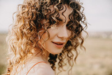 Closeup portrait of a young brunette woman with curly hair, smiling, outdoors, in sunlight.