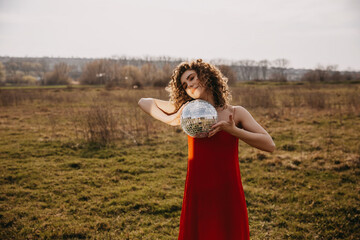 Young woman with curly hair, wearing a red dress, holding a disco ball in a field.