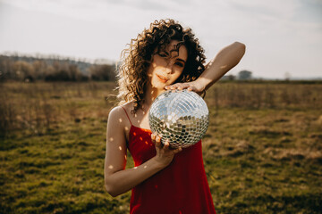 Young woman with curly hair, wearing a red dress, holding a disco ball in sun rays, in a field.