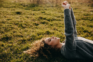 Calm young woman with curly hair, lying on grass, wearing a grey knitted sweater.