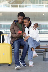 Casual african travelers waiting for taxi at airport terminal use smartphone, watch videos. Travel after end of coronavirus concept. Happy couple, black man and woman arrive on vacation after covid-19
