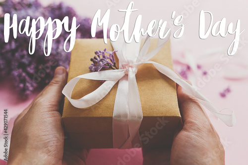 Happy mother's day greeting card. Happy mother's day text and hands giving gift box with ribbon and lilac flowers on pink paper. Stylish floral greetings. Handwritten lettering. Mothers day