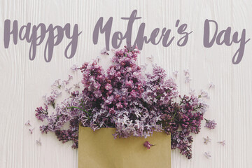 Happy mother's day. Happy mother's day text and purple lilac blooming from craft envelope on white wood flat lay. Stylish floral greeting card. Handwritten lettering. Mothers day
