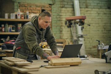 Concentrated adult craftsman checking information on laptop while working in carpentry workshop....