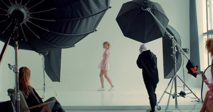 WIDE FIXED Behind the scenes of a photo shoot, team working on a fashion shot, professional female photographer taking pictures of a model in a bright studio. Shot with 2x anamorphic lens
