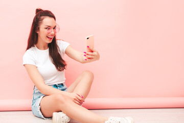Young beautiful smiling female in trendy summer hipster clothes. Sexy carefree woman sitting near pink wall in studio. Positive model having fun indoors. Cheerful and happy. Taking selfie photos