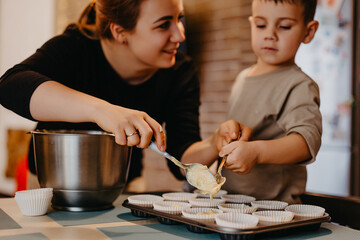 Obraz na płótnie Canvas Mom and her son helping to prepare cupcakes in the kitchen. The boy with plaster on his hand mixes the dough and puts it in baking tins, and mom tells