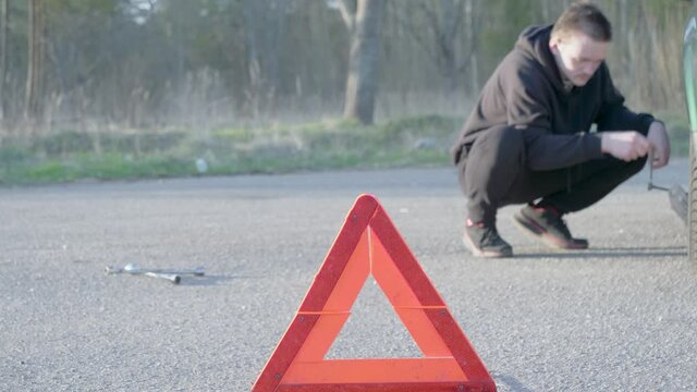 A young guy puts a jack and raises the car, he is wearing a black tracksuit and black sneakers, the concept of a broken car on the road.