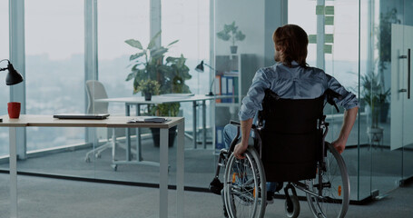 Young caucasian man disabled in wheelchar coming to office corporate workspace sitting by desk...