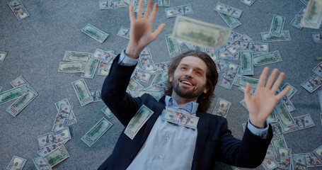 Young happy caucasian office worker lying on the floor cathcing dollar banknotes falling money...