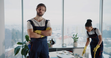 Doiverse corporate team of janitors cleaning corporate office. Indoor portrait of handsome caucasian man cleaner in uniform smiling for camera.