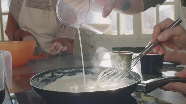 Slow-motion midsection shot of unrecognizable people cooking together making bechamel sauce on frying pan, pouring some cream and whisking it