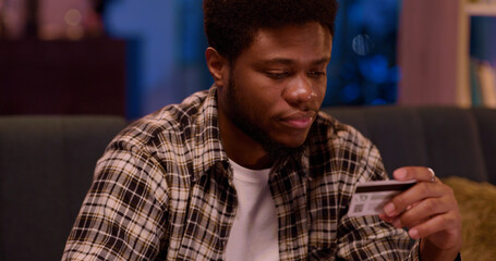 Casual afro-american young man shopping online buying food delivery entering number of credit card using smartphone in living room apartment.