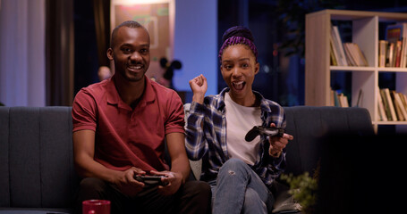 Young african beautiful happy couple playing exciting video game on console using gamepads having fun in living room. Apartment. Entertainment.