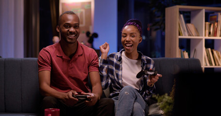 Fototapeta na wymiar Young african beautiful happy couple playing exciting video game on console using gamepads having fun in living room. Apartment. Entertainment.