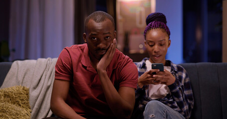 African american love couple at home. Excited male using console gamepad playing online virtual game while his girlfriend using mobile phone.