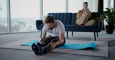 Family stay home. Young woman using tablet relaxing on sofa while her flexible boyfriend man doing head standing yoga pose stretching in living room.