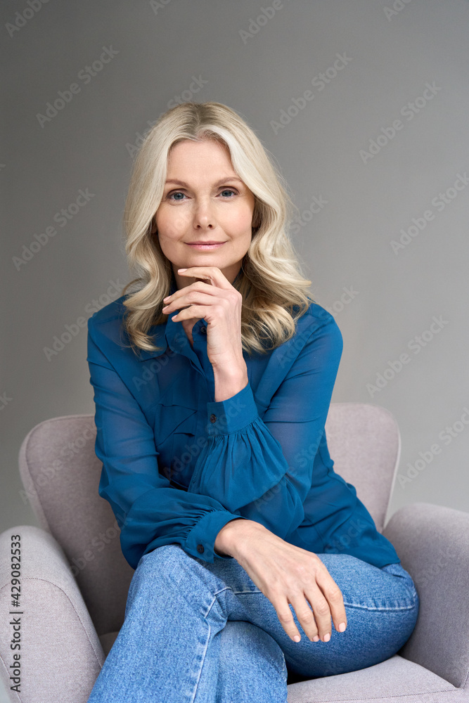 Wall mural Happy smiling adult 50 years old female psychologist sitting on chair looking at camera at gray background. Vertical portrait of sophisticated blonde hair woman advertising products and services.