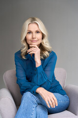 Happy smiling adult 50 years old female psychologist sitting on chair looking at camera at gray...