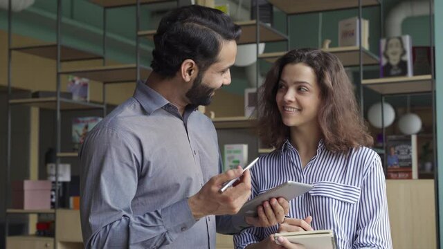 Diverse friendly coworkers talking using digital tablet in office. Indian manager having discussion with latin employee meeting in office lobby discussing project, teaching new employee sharing ideas.
