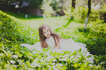 A girl in white dress lies on the grass in spring cherry garden. Portrait of child among white flowers trees. Young lady sitting in sunny blooming park with green grass and yellow dandelions