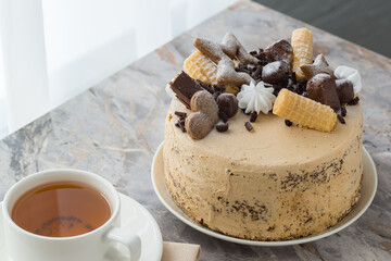 A cup of tea on a saucer and a sponge cake with buttercream on table in the cafe near the window. The cake is decorated with chocolate and cookies