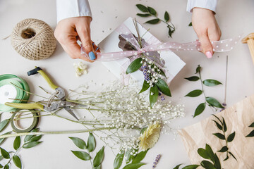 A florist decorates a gift box with flowers and a ribbon on a white desktop. Top view.Only the hands are in the frame