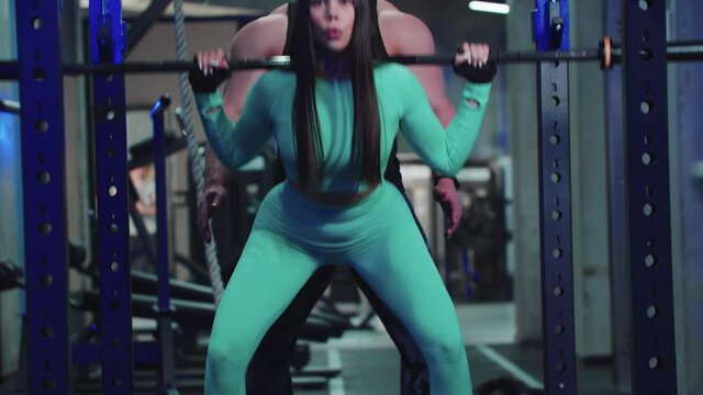 Attactive woman in sports costume trains her butt with dumbbell on her shoulders under the supervision of a trainer 