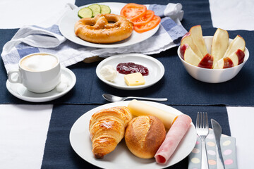 Full breakfast table with croissant, bread, ham and cheese. There are also apple, pretzel, tomato, cucumber, jam, butter, cream and a cup of coffee.