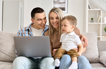 Happy family mother, father and cute child son having fun using laptop at home. Cute toddler kid boy learning computer doing ecommerce online shopping on technology device sitting with parents on sofa