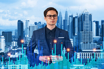 Young eastern handsome businessman pondering about new approaches to consultancy to find priorities for venture capital start up to estimate prospective growth. Financial chart on Kuala Lumpur