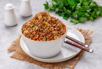 Mushroom Buckwheat Porridge Bowl. Buckwheat Kasha with sauteed mushrooms, onion and carrot served in white ceramic bowl and topped with chopped parsley. Light background, selective focus.